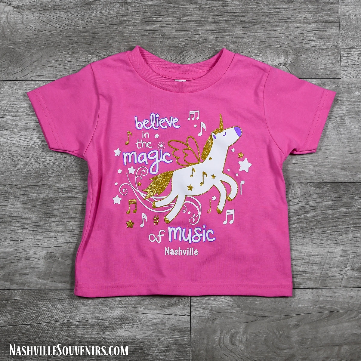 A colorful and sparkly design that reads, "Believe in the Magic of Music Nashville" Toddlers T-Shirt. A Flying horse with gold glitter design and matching stars on a bright pink or purple t-shirt.
