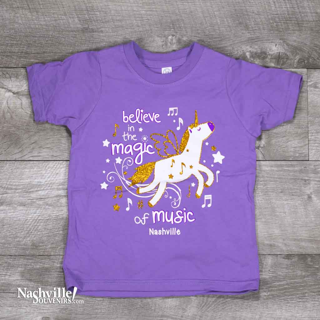 A colorful and sparkly design that reads, "Believe in the Magic of Music Nashville" Toddlers T-Shirt. A Flying horse with gold glitter design and matching stars on a bright pink or purple t-shirt.