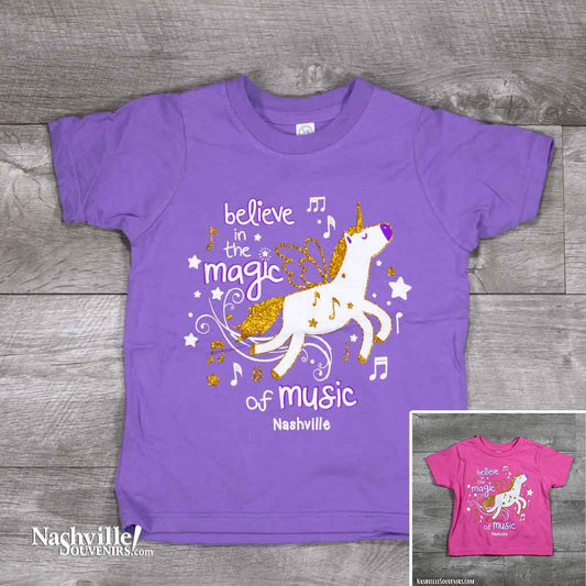 A colorful and sparkly design that reads, "Believe in the Magic of Music Nashville" Toddlers T-Shirt. A Flying horse with gold glitter design and matching stars on a bright pink or purple t-shirt.